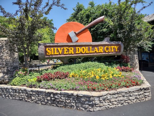 Sign for Silver Dollar City with an axe and vibrant flower beds in front, set against a clear blue sky.