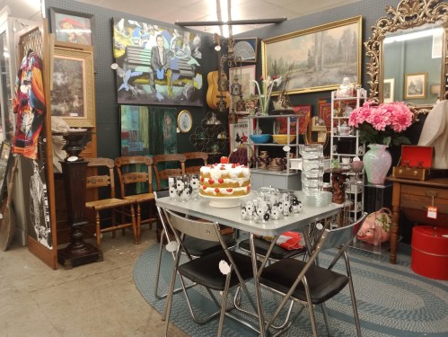 A cozy vintage shop display featuring a table with a cake, chairs, artwork, and various decorative items.
