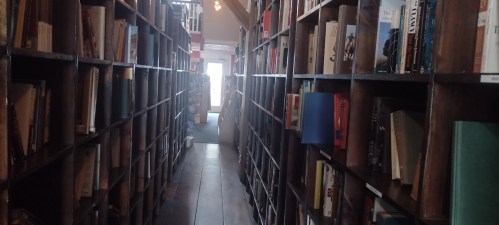 A narrow aisle between tall wooden bookshelves filled with various books, leading to a bright entrance.