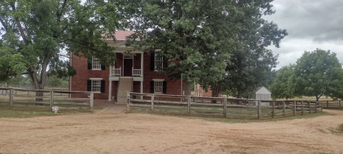 A historic brick building surrounded by trees and a wooden fence, set in a rural landscape with a dirt path.