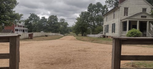 A dirt path leads through a historic site, flanked by trees and old buildings under a cloudy sky.
