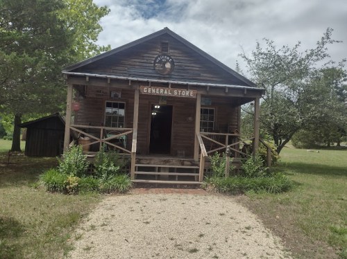 A rustic wooden general store with a porch, surrounded by greenery and a gravel path leading to the entrance.