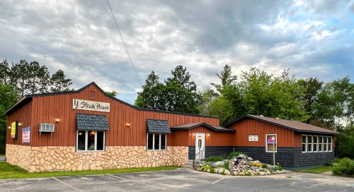 A rustic steakhouse with a wooden exterior, stone accents, and a landscaped entrance under a cloudy sky.