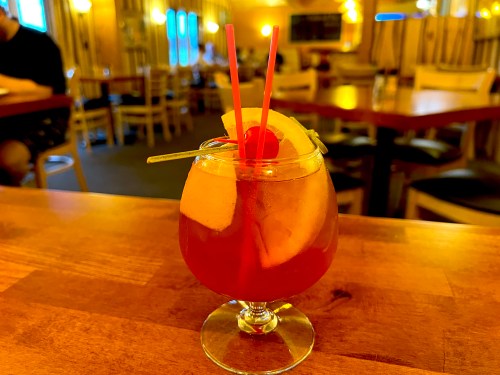 A colorful cocktail in a glass with a cherry and fruit garnish, served on a wooden table in a cozy restaurant.