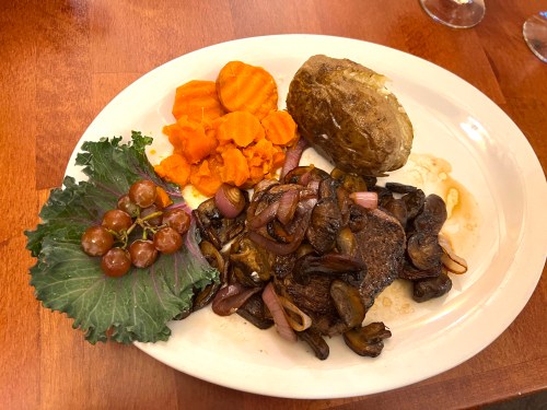 A plate featuring steak with mushrooms and onions, sweet potatoes, a baked potato, and a side of grapes and greens.