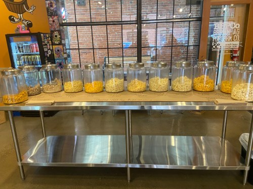 A display of various popcorn flavors in glass jars on a stainless steel table, with a colorful background.