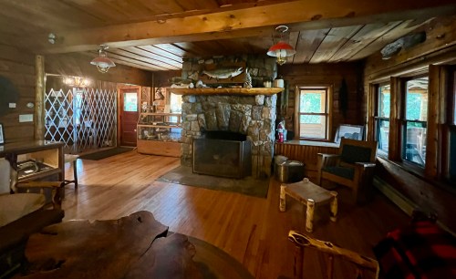 Cozy log cabin interior with wooden floors, stone fireplace, and rustic furniture, featuring large windows and warm lighting.