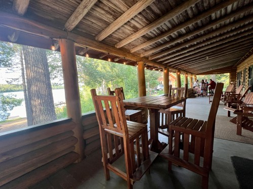 A wooden porch with a table and chairs overlooking a lake, surrounded by trees and a cozy cabin atmosphere.