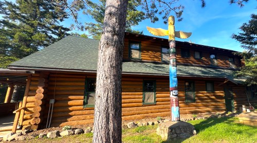 A log cabin with a colorful totem pole in front, surrounded by trees and greenery.