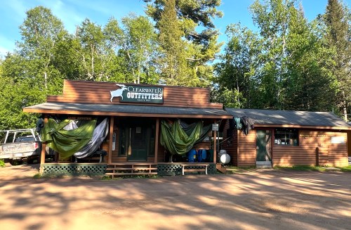 A rustic building with a sign reading "Clearwater Outfitters," surrounded by trees and outdoor gear.