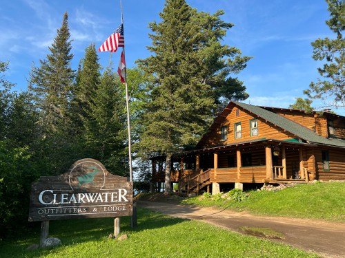 A log cabin lodge surrounded by trees, with a sign reading "Clearwater Outfitters & Lodge" and an American flag.