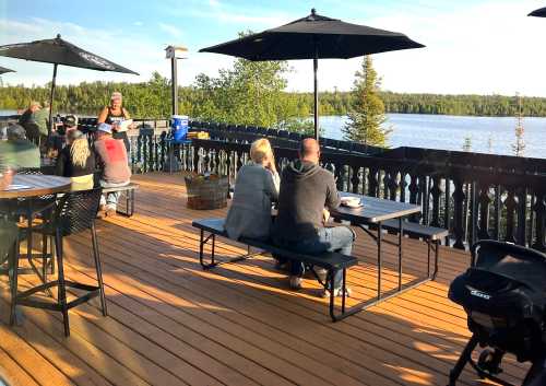 A lakeside patio with people dining, enjoying the view, and a server attending to customers under umbrellas.