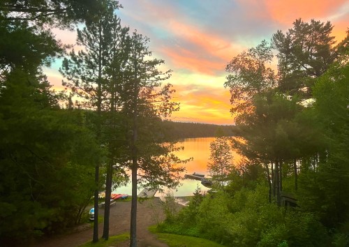 A serene lake at sunset, surrounded by trees, with colorful skies reflecting on the water and boats along the shore.