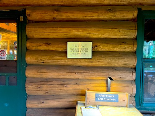 Sign on a log cabin wall indicating it's on the National Register of Historic Places, with a self-check-in station below.