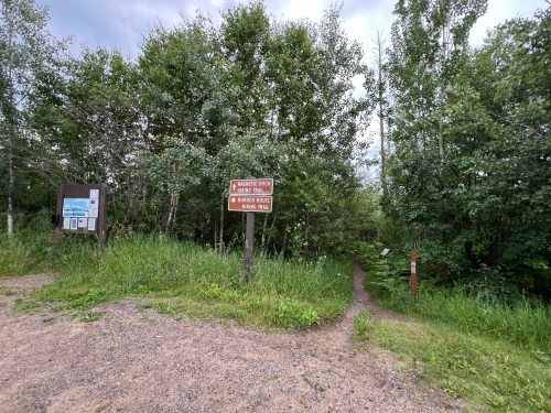 Trail intersection with signs for "Murder Rock" and "Bridger Mountain Hiking Trail," surrounded by greenery.