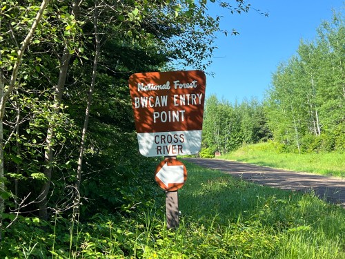 Sign for the Boundary Waters Canoe Area Wilderness (BWCAW) entry point at Cross River, surrounded by trees and grass.