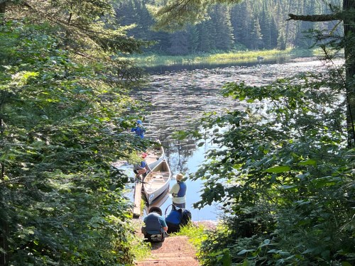 A serene lakeside scene with people preparing a canoe amidst lush greenery and calm water.