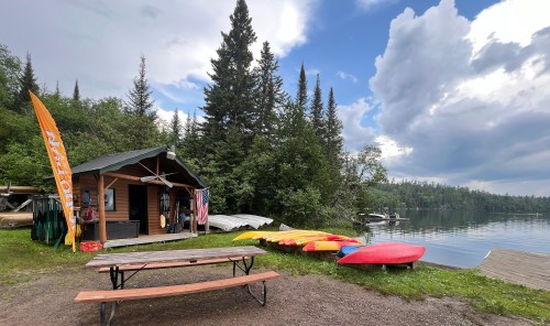 A lakeside cabin with kayaks, a picnic table, and trees under a cloudy sky.