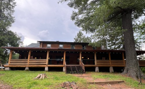 A large log cabin with a spacious porch, surrounded by trees and grass, under a cloudy sky.