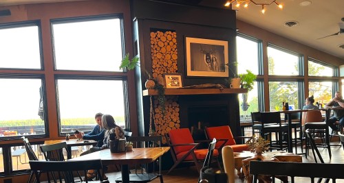 Cozy restaurant interior with large windows, wooden decor, and patrons enjoying their meals at tables.