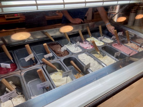 A display of various ice cream flavors in metal tubs, with wooden scoops and labels for each flavor.