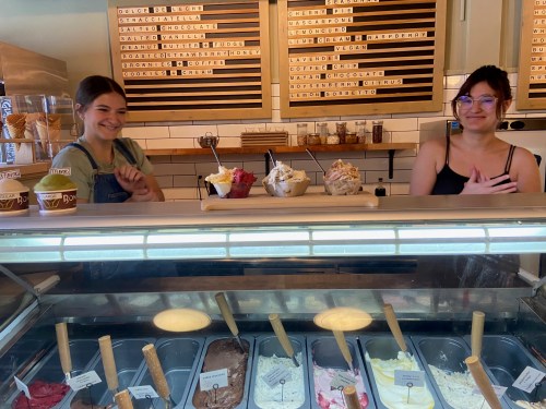 Two smiling employees stand behind a gelato counter, showcasing various flavors in tubs and serving cups.