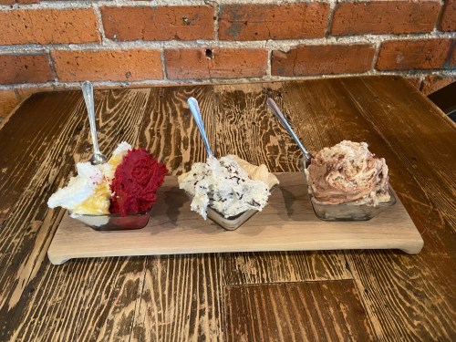 Three bowls of ice cream on a wooden board: red velvet, cookies and cream, and chocolate fudge, with spoons.