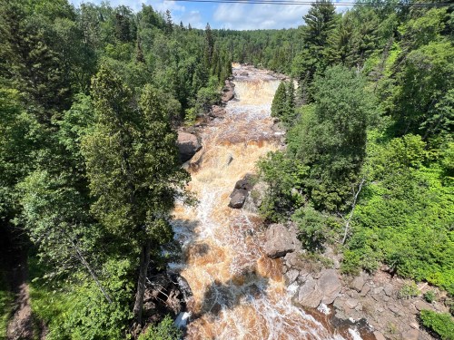 A cascading waterfall flows through a lush green forest, surrounded by trees and rocky terrain under a blue sky.