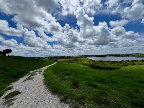 A winding gravel path leads through lush green fields under a bright blue sky filled with fluffy white clouds.