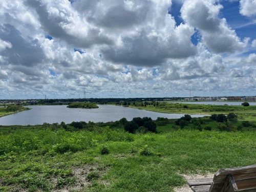 A scenic view of a lake surrounded by greenery and clouds in a blue sky.