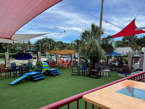 Outdoor recreational area with colorful seating, shaded by canopies, and a grassy surface, surrounded by palm trees.