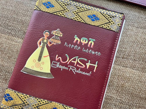 A colorful menu cover for WASH Ethiopian Restaurant featuring traditional designs and an illustration of a woman in cultural attire.