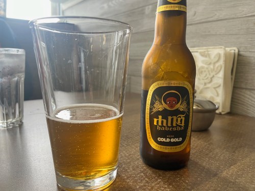 A glass of golden beer next to a bottle labeled "Habesha Gold Gold" on a wooden table.