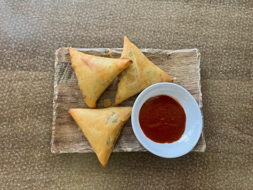 Three golden-brown samosas on a wooden platter, accompanied by a small bowl of red dipping sauce.