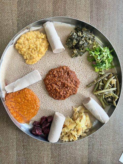 A round platter with injera topped with various colorful Ethiopian dishes, including lentils, greens, and vegetables.