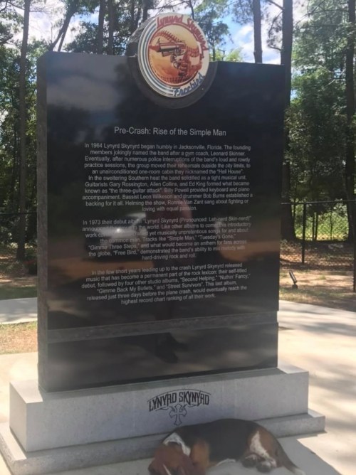 A memorial stone for Lynyrd Skynyrd, detailing the band's history, with a dog resting nearby in a wooded area.