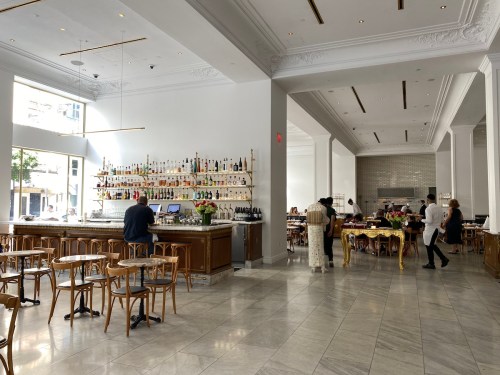 A spacious, modern restaurant interior with a bar, shelves of bottles, and patrons dining at wooden tables.