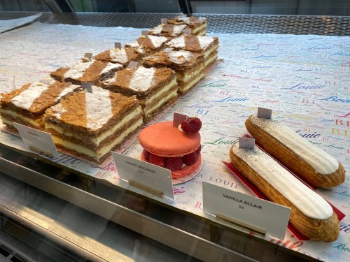 A display of pastries including layered cakes, a macaron, and a vanilla éclair on a colorful patterned surface.
