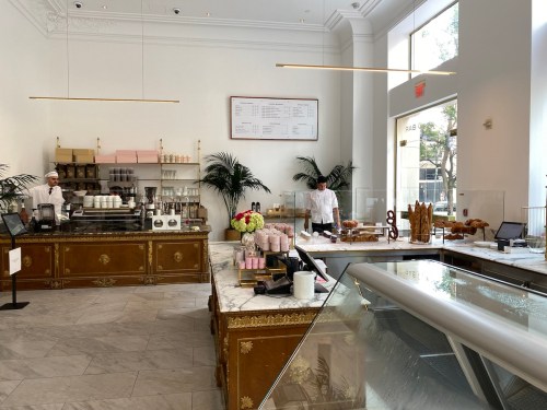 A bright, elegant café interior with a display of pastries, a counter, and two staff members preparing food.