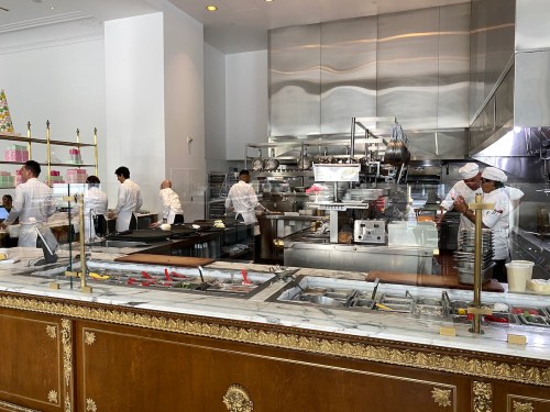 A busy kitchen with chefs preparing food, featuring stainless steel equipment and a marble countertop.
