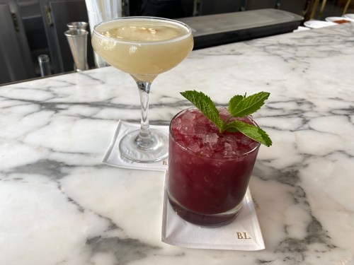 Two cocktails on a marble countertop: a pale drink in a coupe glass and a dark red drink with mint in a rocks glass.