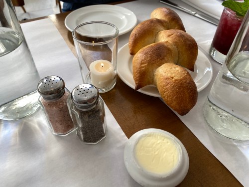 A plate of golden-brown rolls next to a candle, salt and pepper shakers, and a dish of butter on a wooden table.