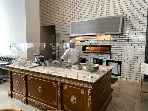 A chef in a white uniform prepares food at a marble countertop in a modern kitchen with a brick wall and oven.