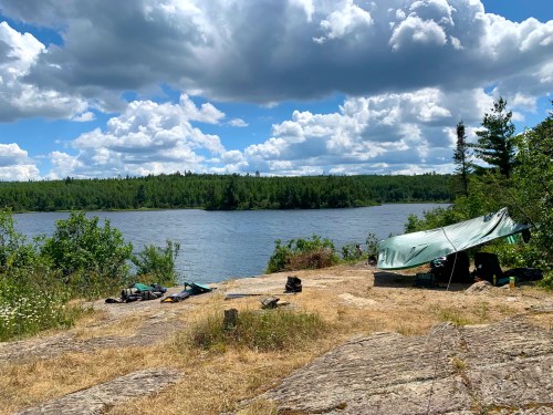A serene lakeside campsite with a green tarp, surrounded by trees and a blue sky filled with fluffy clouds.