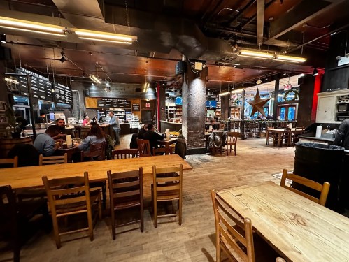 A cozy restaurant interior with wooden tables, chairs, and a bustling counter area, featuring warm lighting and decor.
