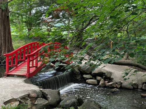 A red wooden bridge arches over a small waterfall, surrounded by lush green trees and rocks.