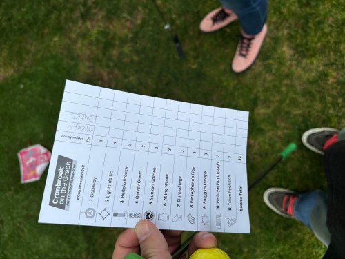 A hand holds a scorecard for a mini-golf game, with a green lawn and feet in the background.