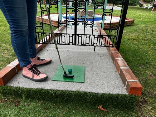 A person in pink shoes prepares to putt a golf ball on a green mat near a decorative fence in a grassy area.