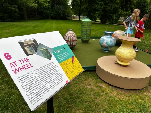 A sign titled "At the Wheel" next to colorful pottery sculptures on a mini-golf course, with children playing nearby.