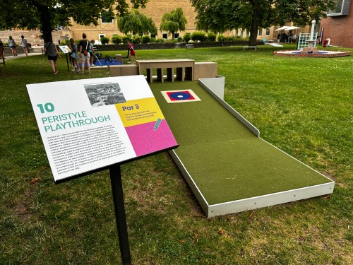 A grassy area featuring a sign about "Peristyle Playthrough" and a sloped play structure with a target.